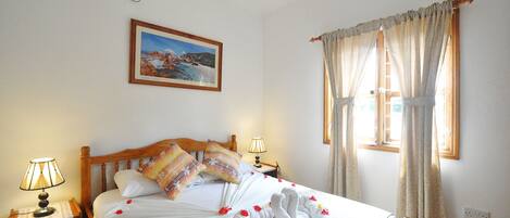 Una cama cuidadosamente arreglada con pétalos de rosa y toallas, un cabecero de madera, una mesita de noche con lámpara, un cuadro en la pared y una ventana con cortinas.