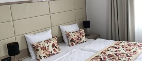 A hotel room with a bed, two nightstands, a lamp, a floral bedspread, and a view of the outside through a window with curtains.