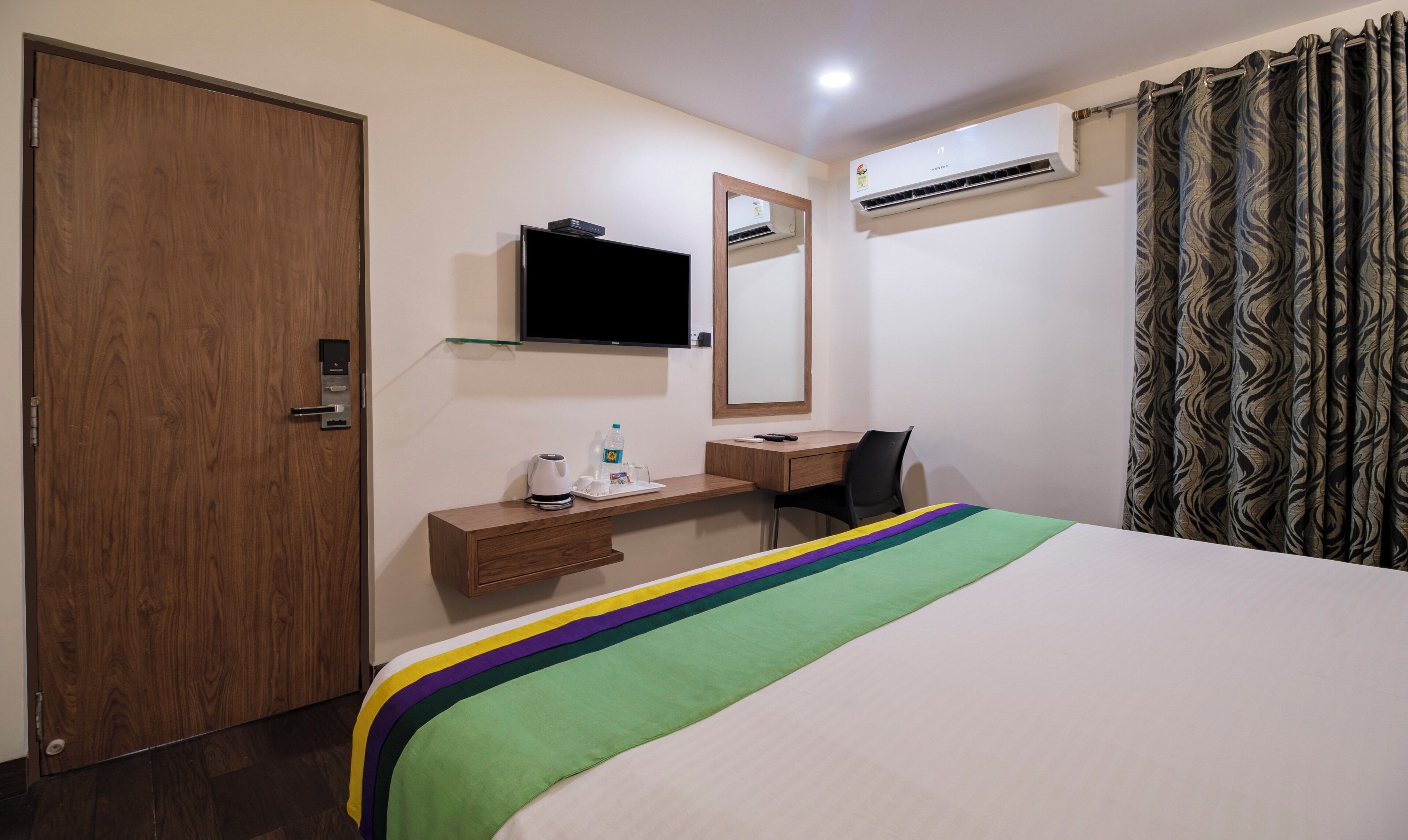 Une chambre d’hôtel avec une porte en bois, un bureau avec une chaise, un lit recouvert d’une couverture colorée, un téléviseur fixé au mur et un appareil de climatisation.