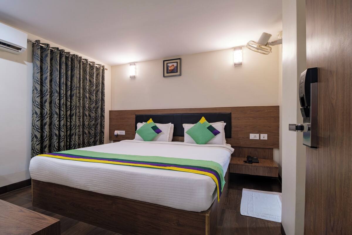 A hotel room with a bed, a nightstand, an air conditioning unit, a window with curtains, and a door.