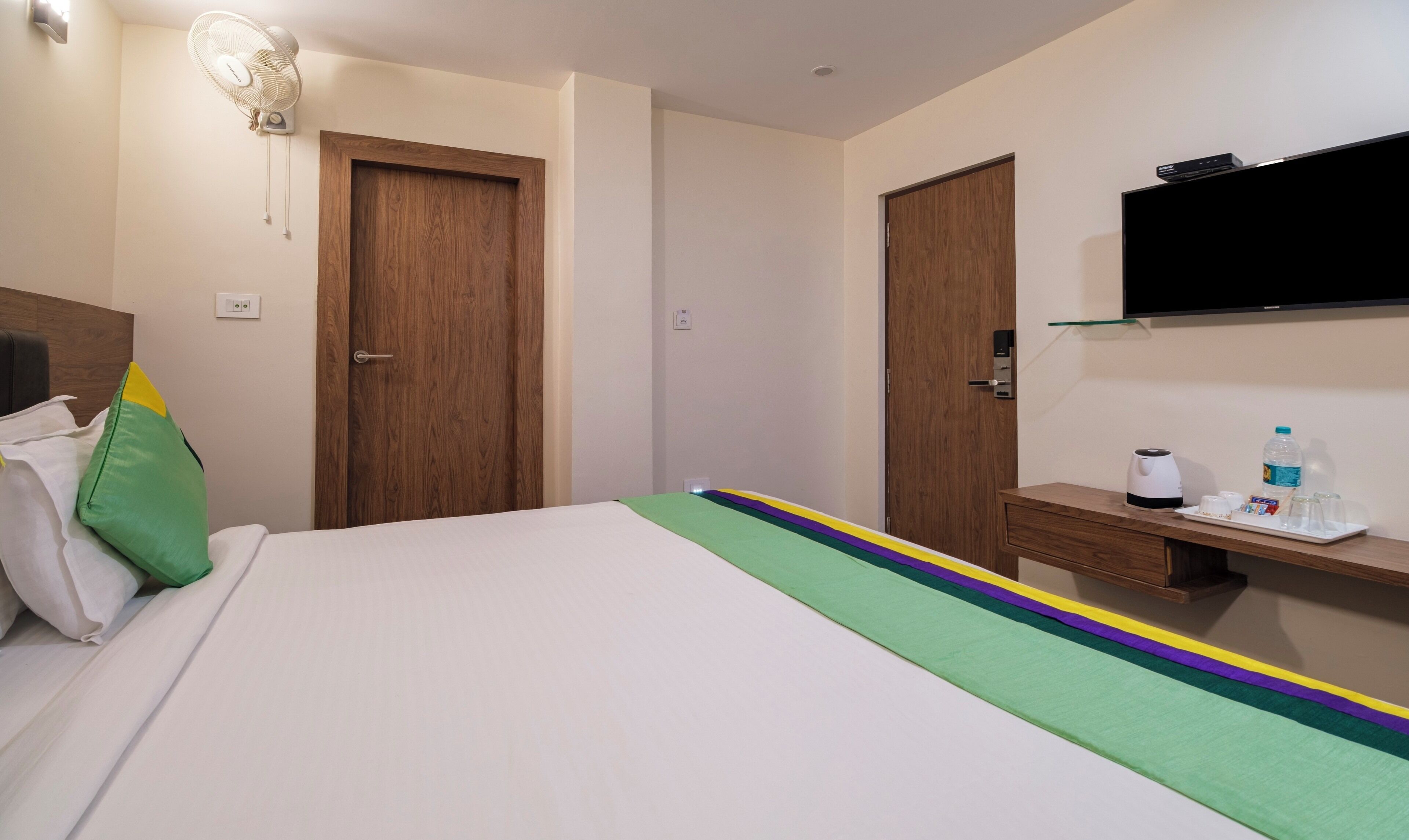 A hotel room with a bed, a bedside table with a kettle, a water bottle, and glasses. There are two wooden doors and a flat-screen TV mounted on the wall.