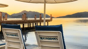 Dos sillones blancos con cojines azules y el nombre "KAYAMARIS" en los respaldos, ubicados bajo un parasol junto a un cuerpo de agua tranquilo con montañas al fondo.