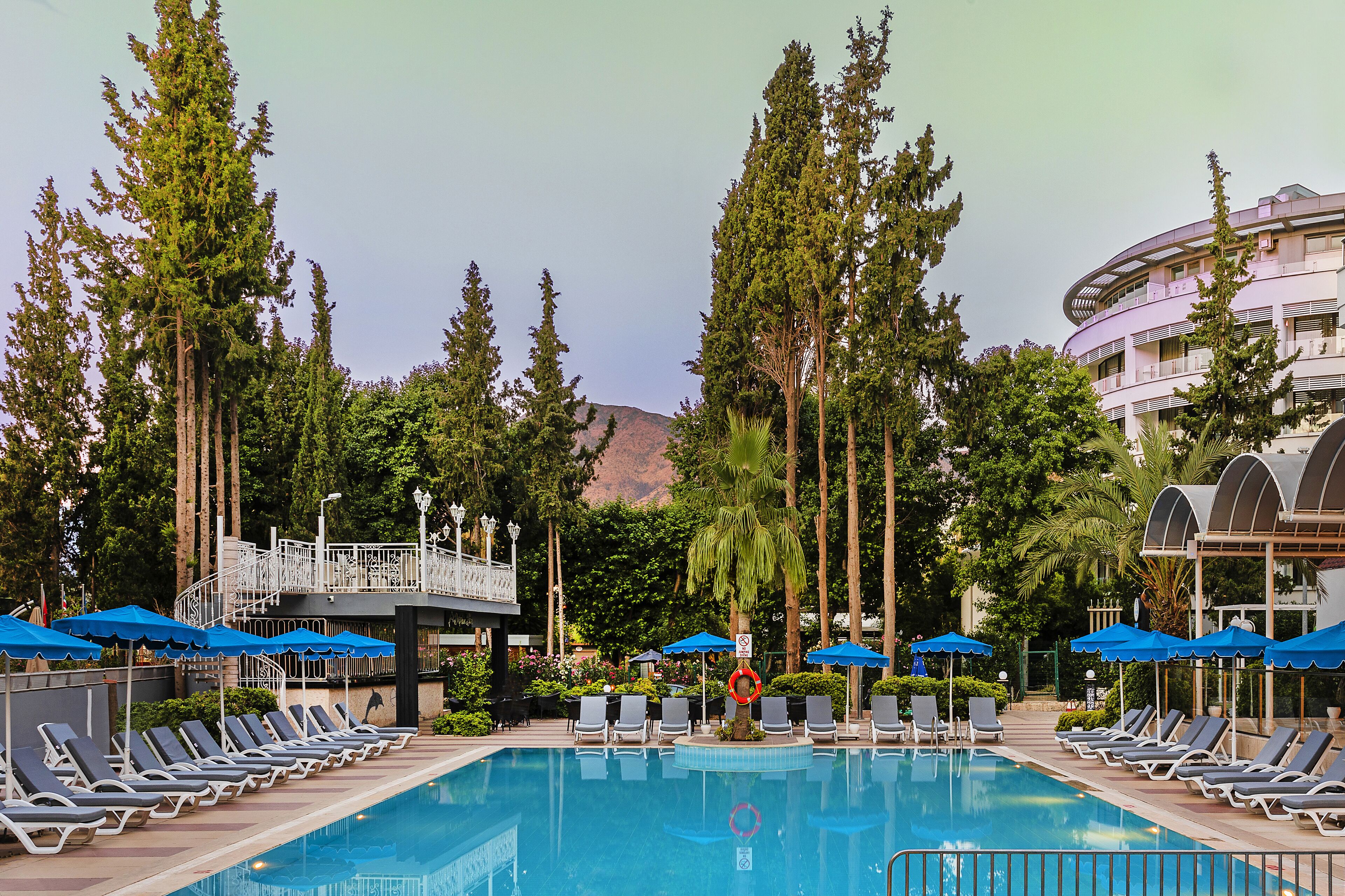 A pool area with lounge chairs and umbrellas, surrounded by tall trees and a building with a curved balcony.