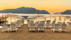 Una playa con filas de sillas reclinables blancas y sombrillas, con la etiqueta "Kayamars", y vista a montañas al fondo.