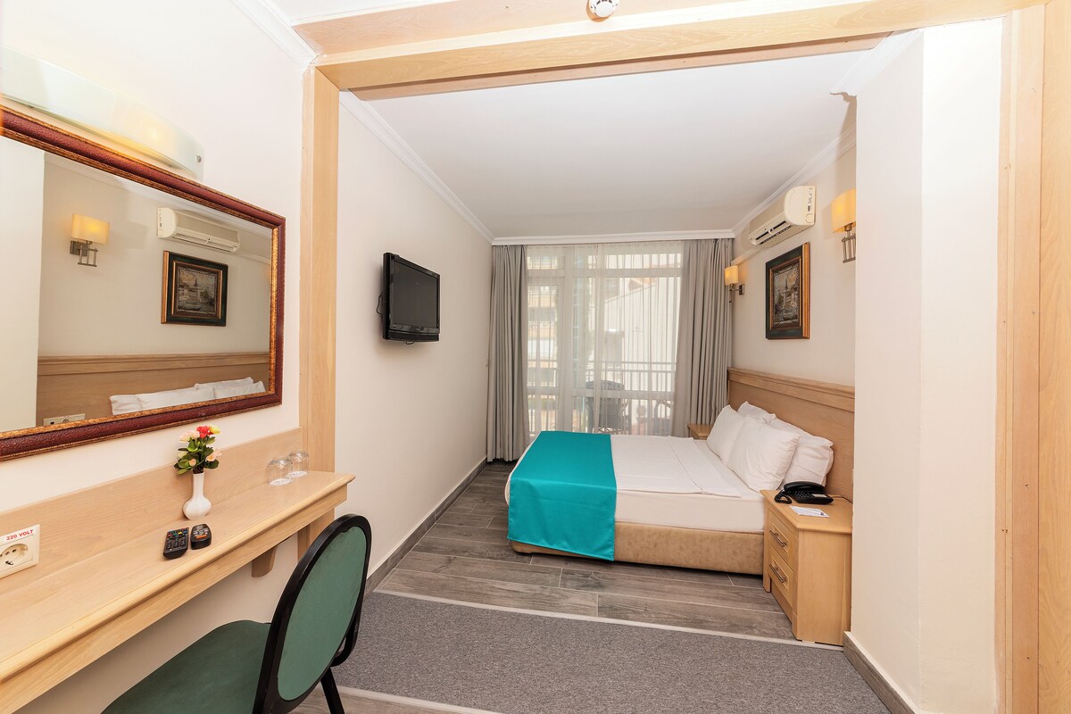 A hotel room with a bed, a desk with a chair, a TV, and a window with curtains.