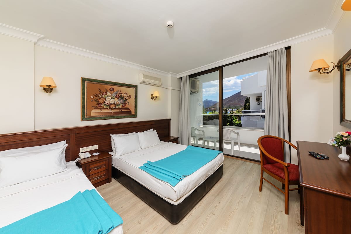 A hotel room with two beds, a desk, and a chair. There is a painting on the wall and a window with a view of the mountains.