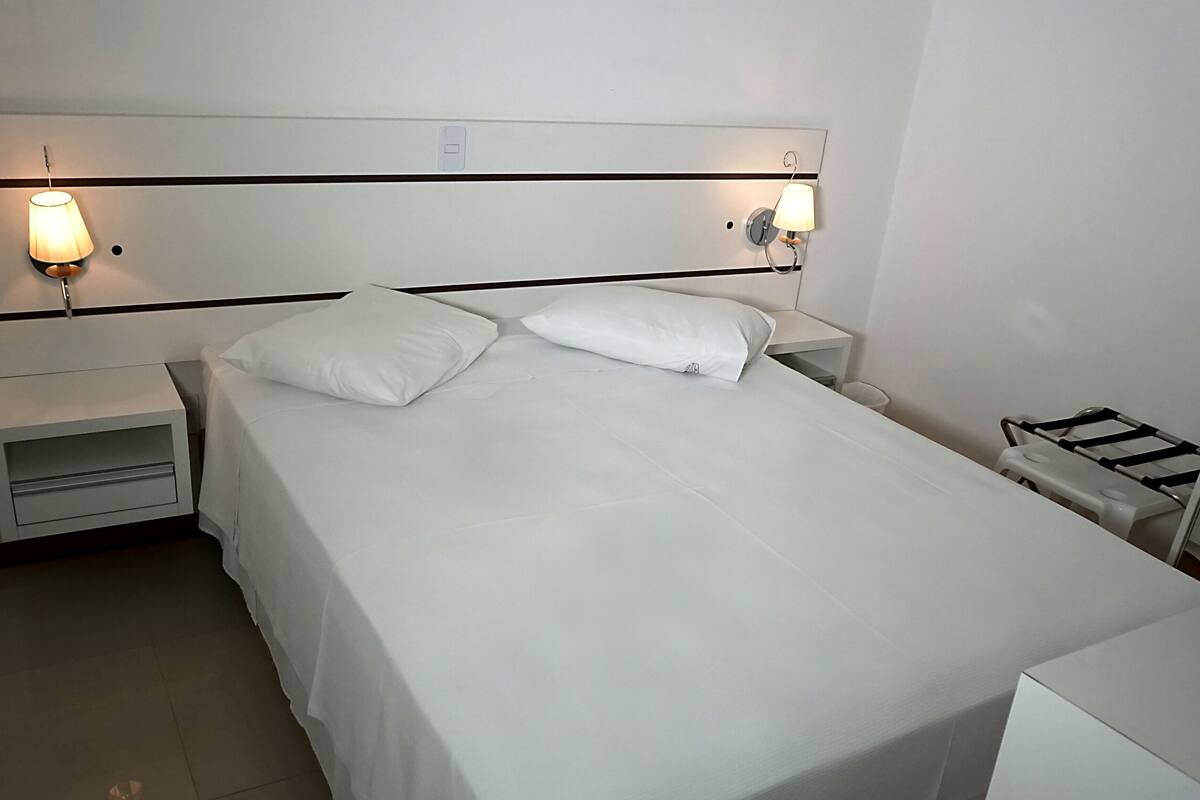 A neatly made bed with white linens and two pillows, flanked by bedside tables with lamps.