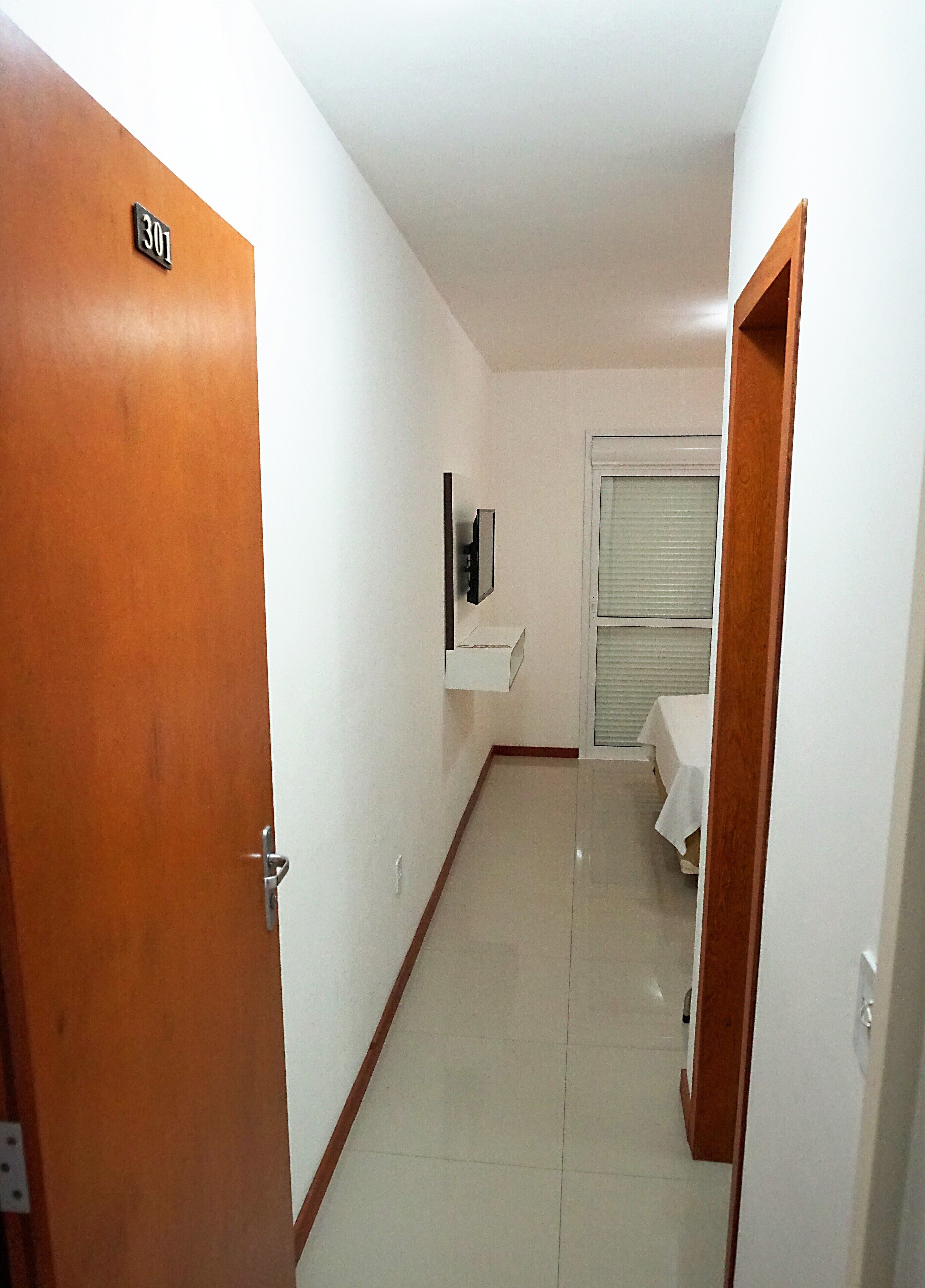 A hallway with a wooden door labeled "301", a white wall-mounted TV, a window with blinds, and a small table with a white tablecloth.