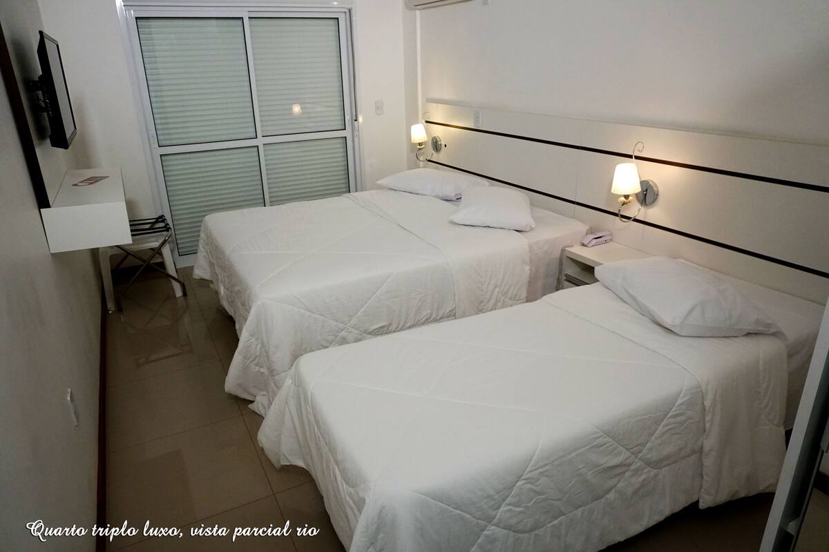 A room with two single beds, a bedside table with a lamp, a TV mounted on the wall, and a window with blinds.