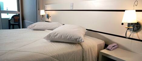 Una habitación de hotel con una cama grande, dos lámparas de noche, una mesita con un teléfono y una silla junto a la ventana.