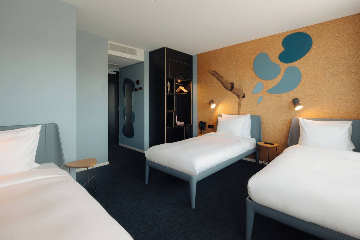 Quarto de hotel com duas camas, uma mesa pequena, um guarda-roupa e uma parede com arte abstrata.