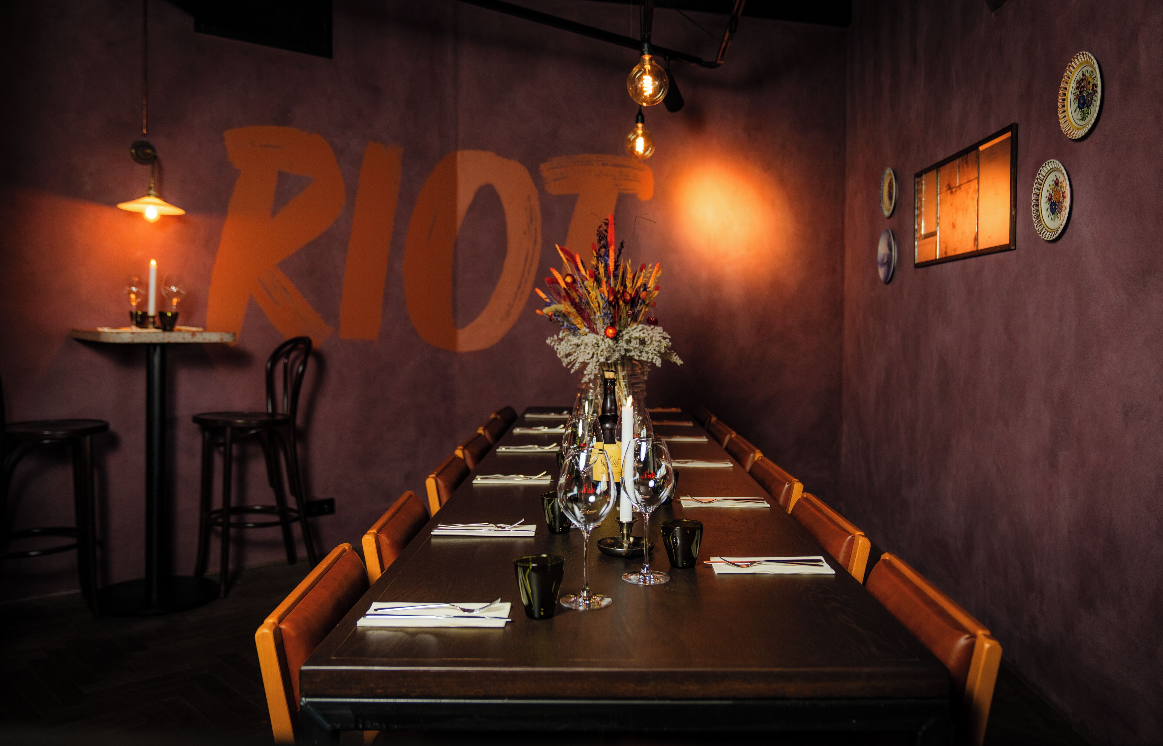 A restaurant with a long table set for dining, a vase with flowers, and a wall with the word "RIOT" in large letters.