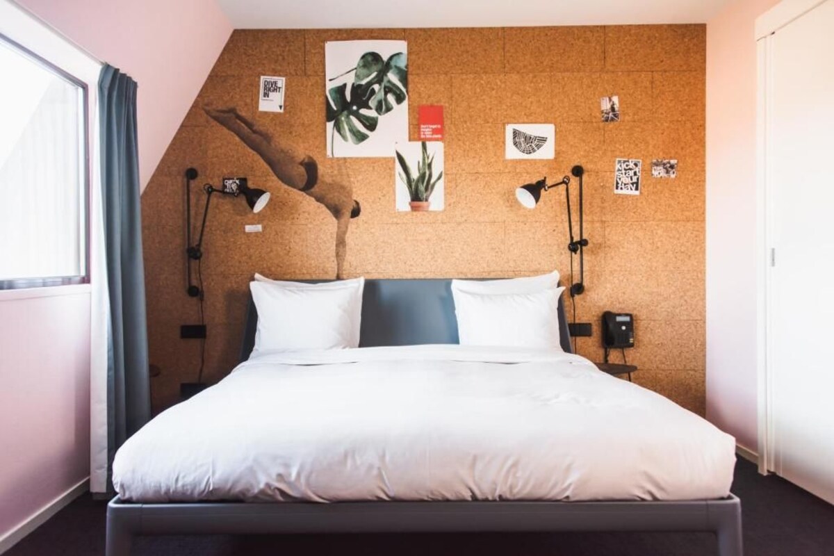 A neatly made bed in a room with a corkboard wall adorned with various items, including a plant and artwork.