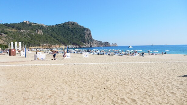 Una playa con tumbonas y sombrillas, una red de voleibol y una colina rocosa al fondo.