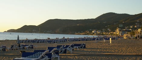 A beach with rows of sun loungers and umbrellas, a clear sky, and a hilly landscape in the background.