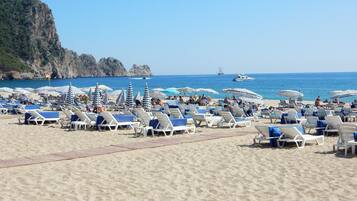 A beach with rows of sun loungers and umbrellas, a rocky cliff in the background, and a boat in the water.