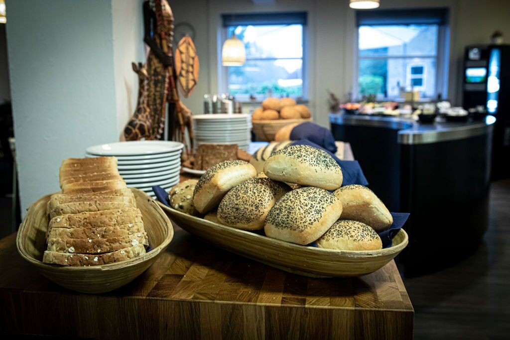 En buffetbord med forskellige brød og kager, herunder valnødderunderlagte rundstykker og skiveskåret brød, i et godt oplyst rum med udsigt over det fri gennem store vinduer.