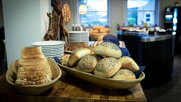 En buffetbord med forskellige brød og kager, herunder valnødderunderlagte rundstykker og skiveskåret brød, i et godt oplyst rum med udsigt over det fri gennem store vinduer.