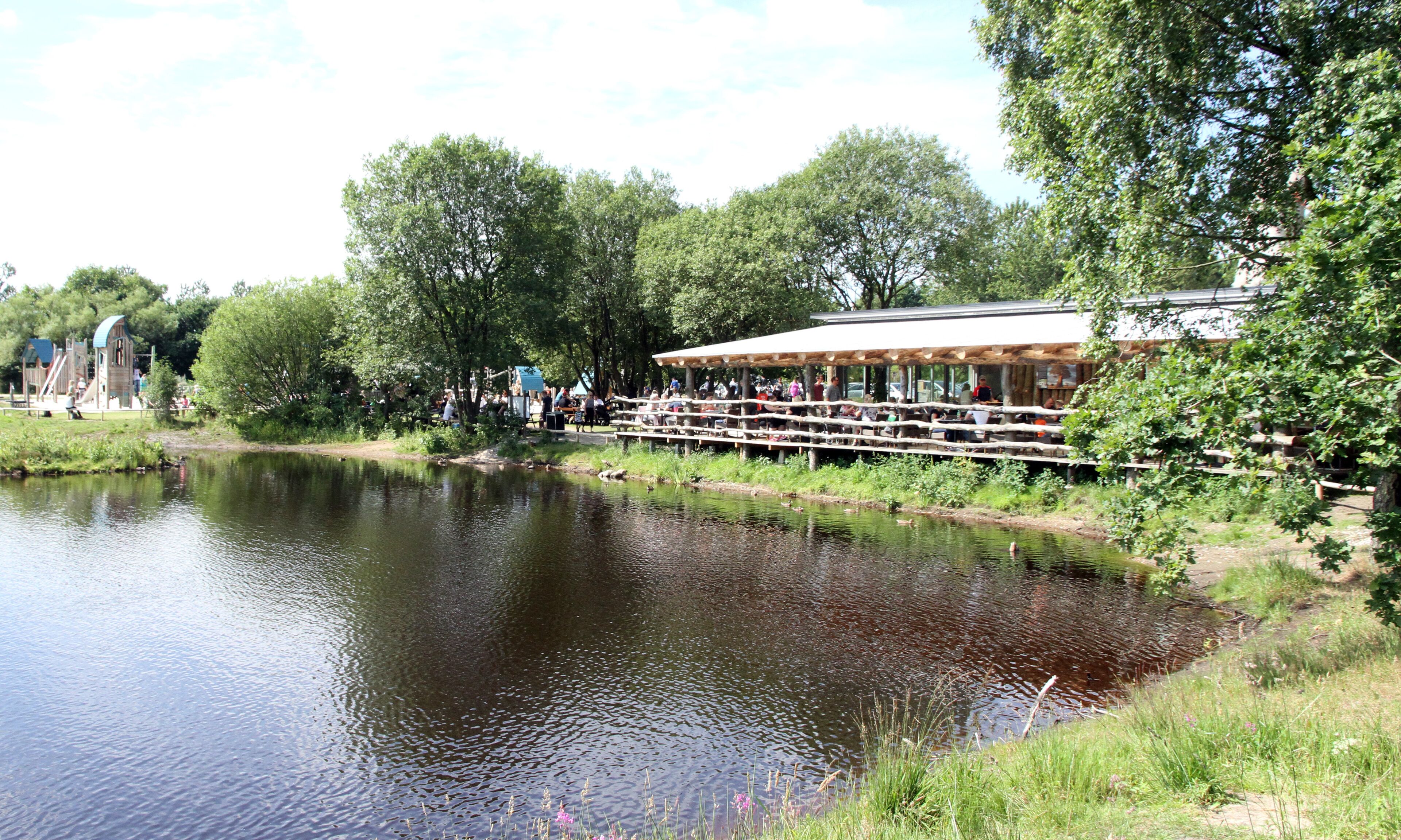 A riverside restaurant with outdoor seating overlooking a calm river, surrounded by lush greenery.