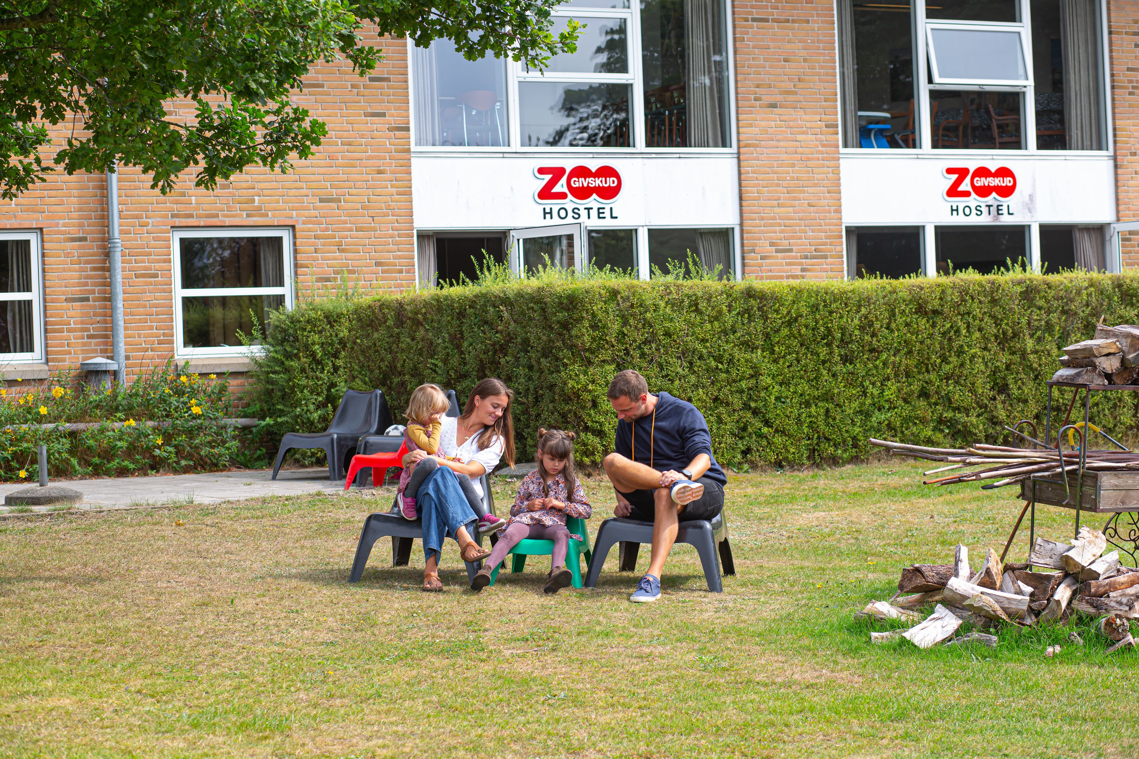 A family sitting on plastic chairs outdoors in front of a building with a sign reading "ZGVSKUD HOSTEL".