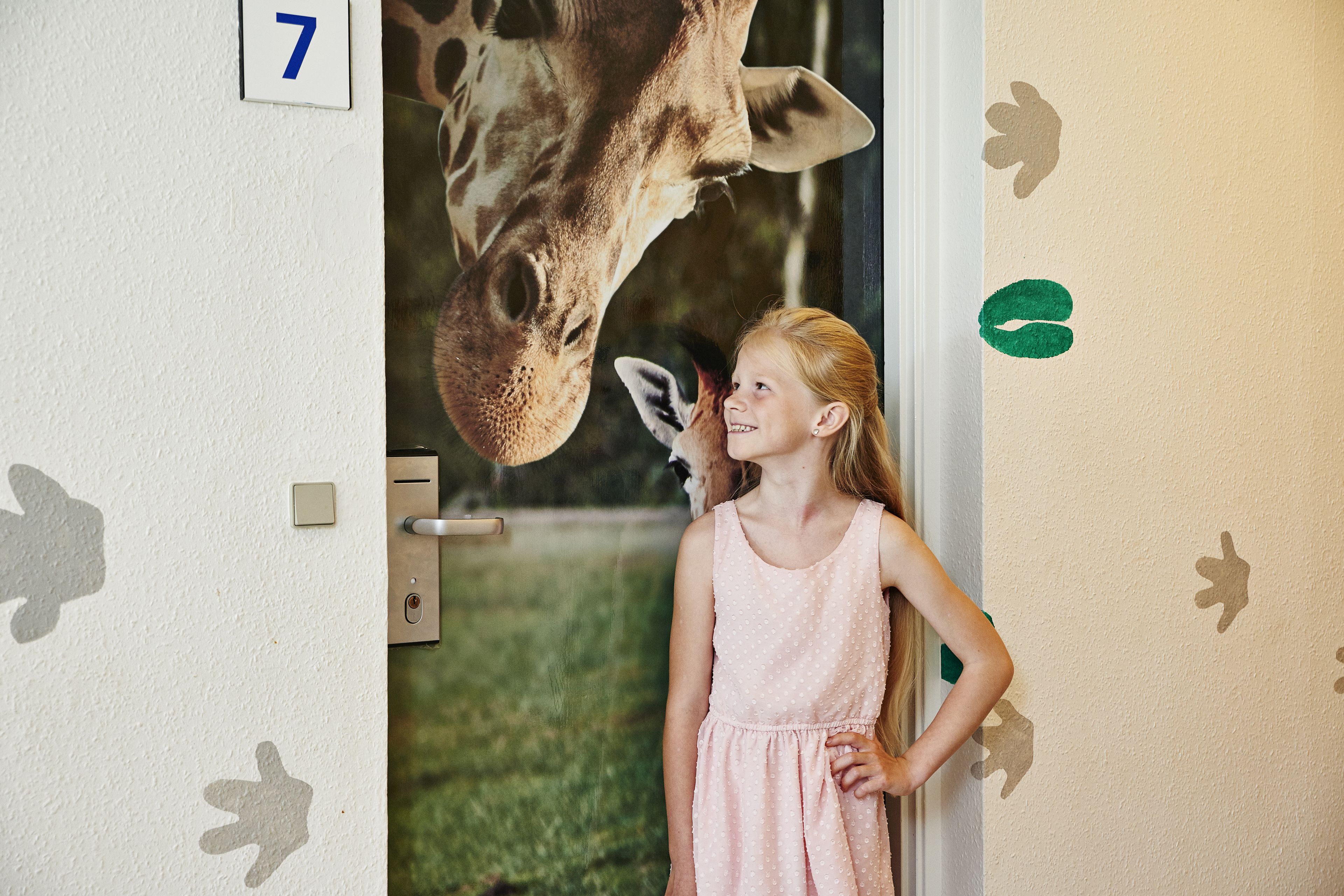 A room with a door marked with the number 7, a giraffe head peering through the door, and a child standing by the door.