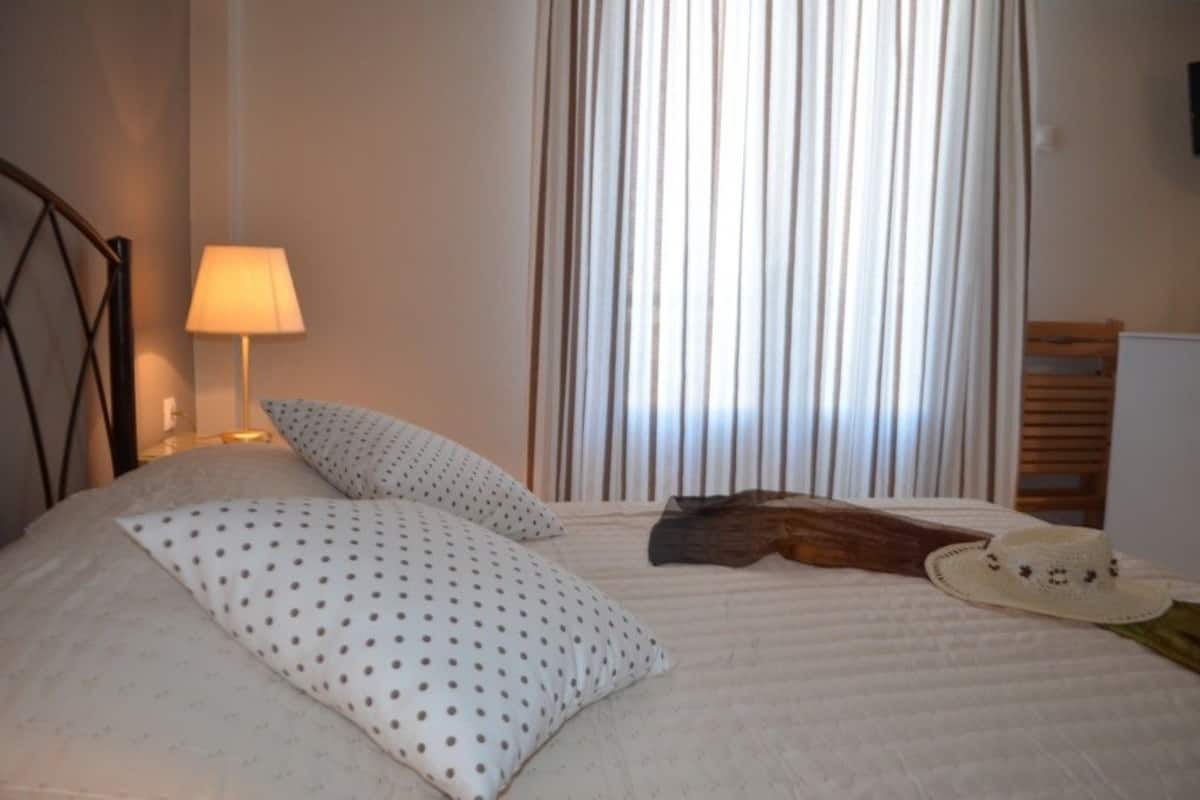 A neatly made bed with white bedding and polka dot pillows, a wooden headboard, a bedside lamp, and a hat on the bed.