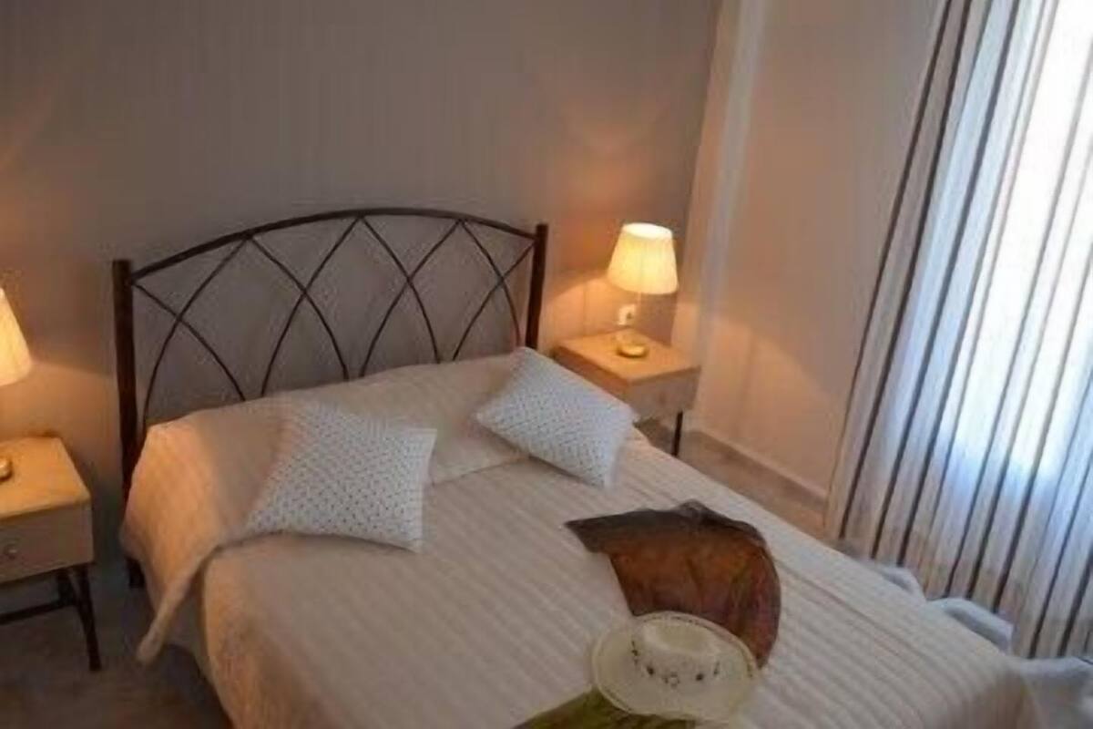 A neatly made bed with a headboard, two pillows, a bedside lamp, and a hat on the bed.