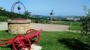 A stone structure with a metal pot on top, a red carriage, and a view of a town in the background. - Capanna 1826 (San Gimignano)