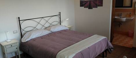 A single bed with a headboard, two bedside tables, a wall-mounted artwork, and a bathroom with a sink and mirror.