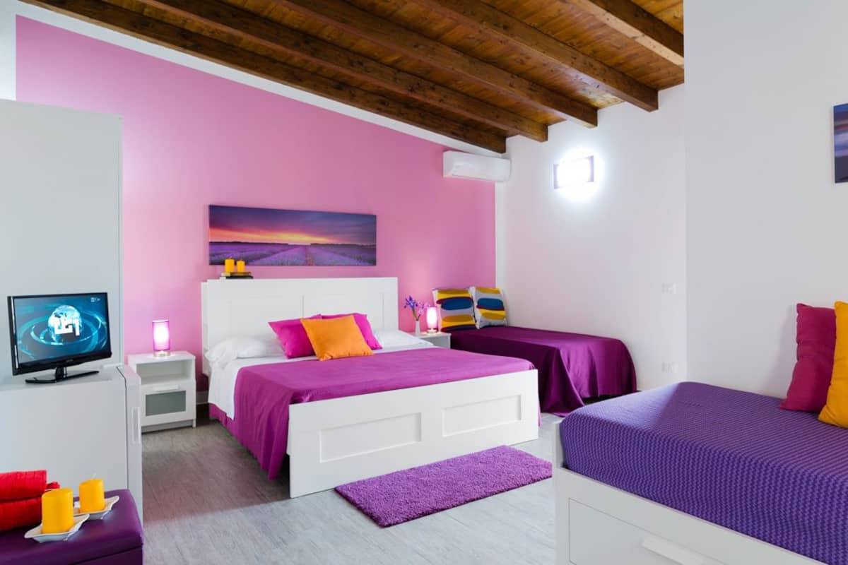 Ein Schlafzimmer mit pinkfarbener Wand, einem Bett mit violett-orange gestreuter Bettwäsche, einem Fernseher und einem Sonnenuntergangsbild.