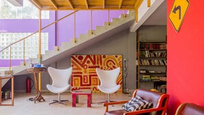 A modern living room with a staircase, a bookshelf, and a vibrant abstract painting. - Casa Bacarirá (São Sebastião)