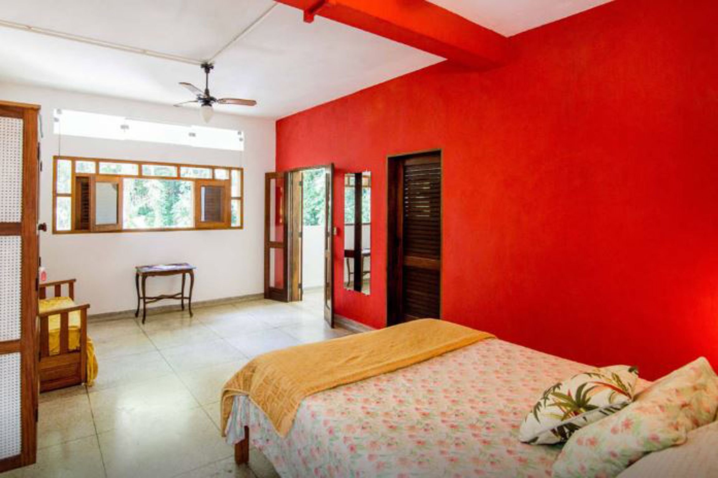 A bedroom with a red wall, a bed with floral bedding, a wooden chair, a small table, and a window with a view of greenery.