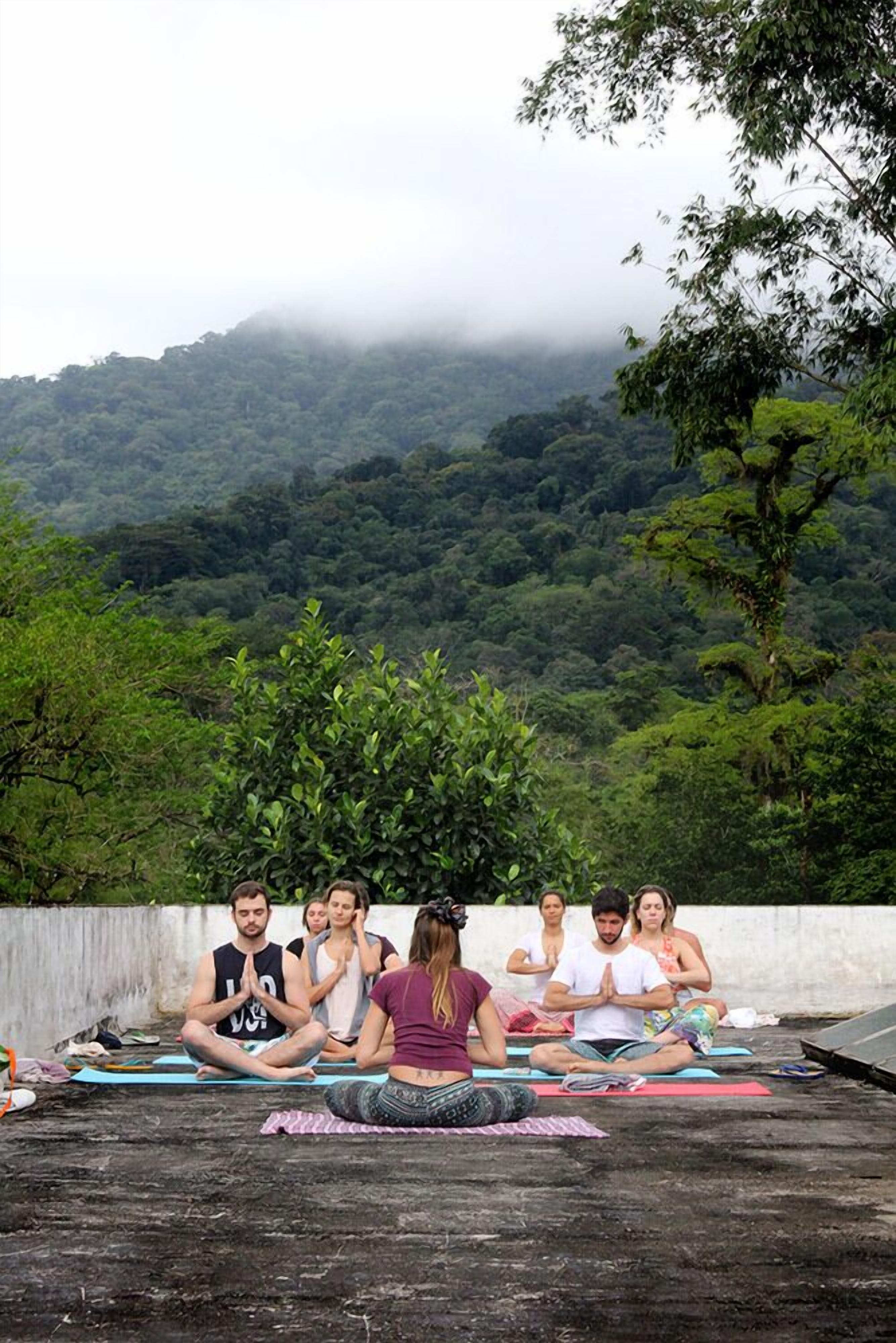 Um grupo de pessoas meditando em tapetes de yoga numa plataforma ao ar livre, com uma montanha arborizada ao fundo.