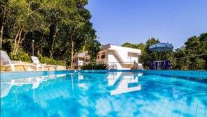 A swimming pool with clear blue water, surrounded by greenery and a white building in the background. - Casa Bacarirá (São Sebastião)