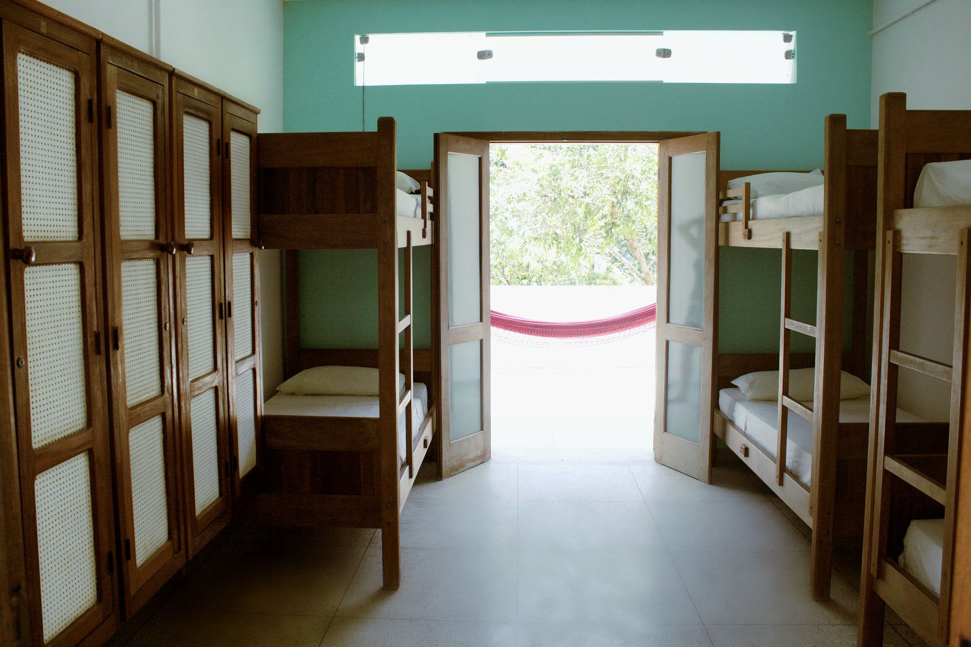 A dormitory room with bunk beds, a window with a view of greenery, and a door leading outside.