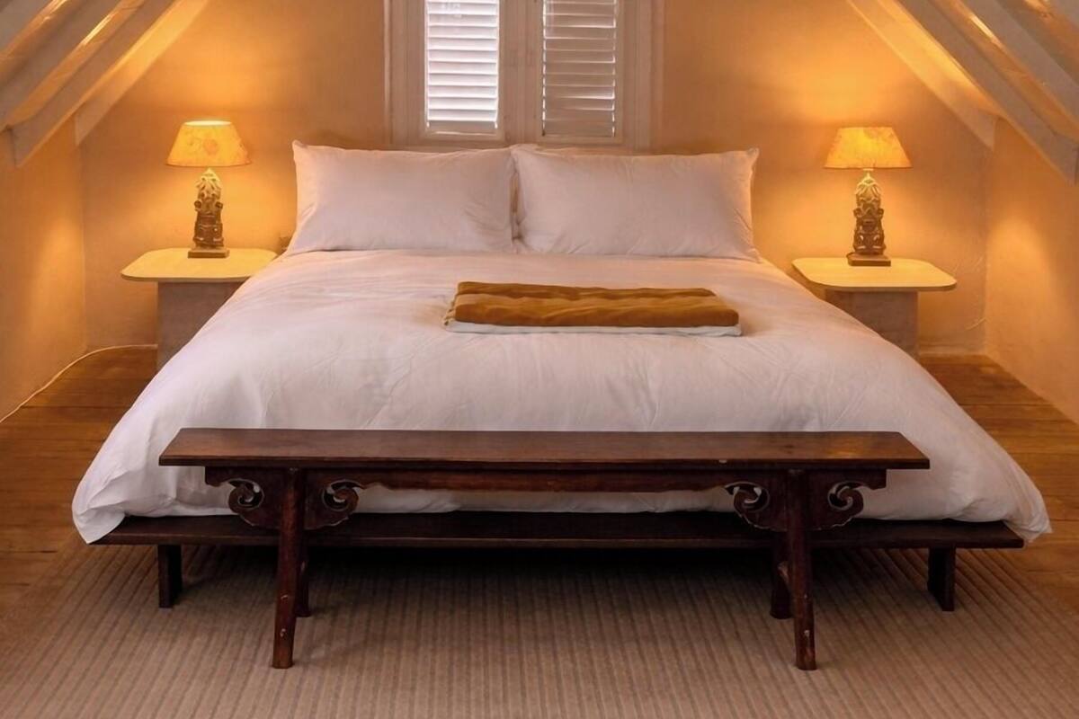 A bedroom with a bed, two bedside tables with lamps, a window with shutters, and a sloped ceiling with exposed beams.