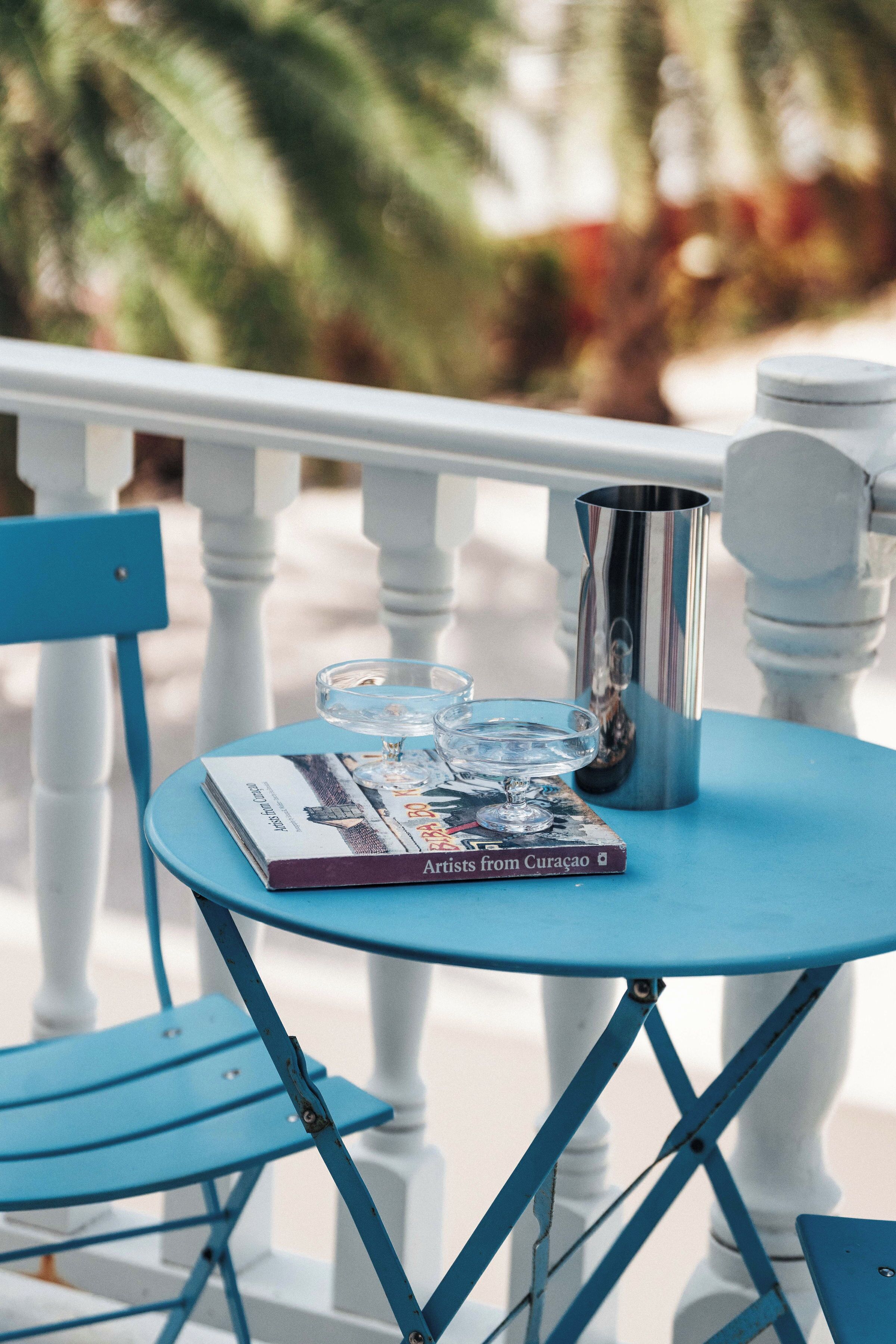 Una mesa azul con una revista, un vaso y una jarrón metálico en un balcón con barandas blancas.