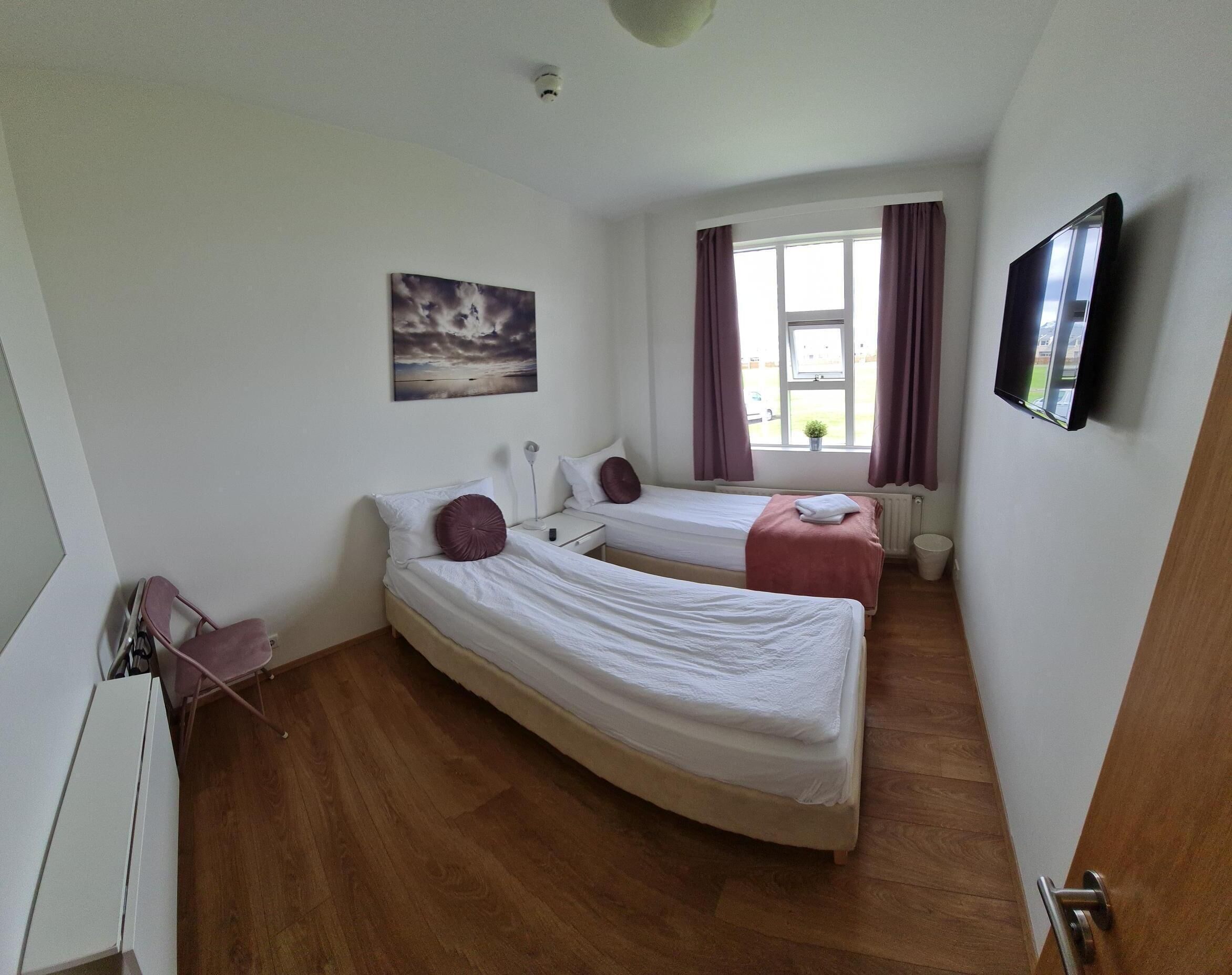 A small, clean room with two single beds, a window with curtains, a mounted TV, and a ceiling-mounted security camera.