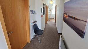 A hallway with a wooden door, a black chair, a bulletin board with notices, a radiator, and a large landscape painting on the wall. - Höfn guesthouse (Höfn)