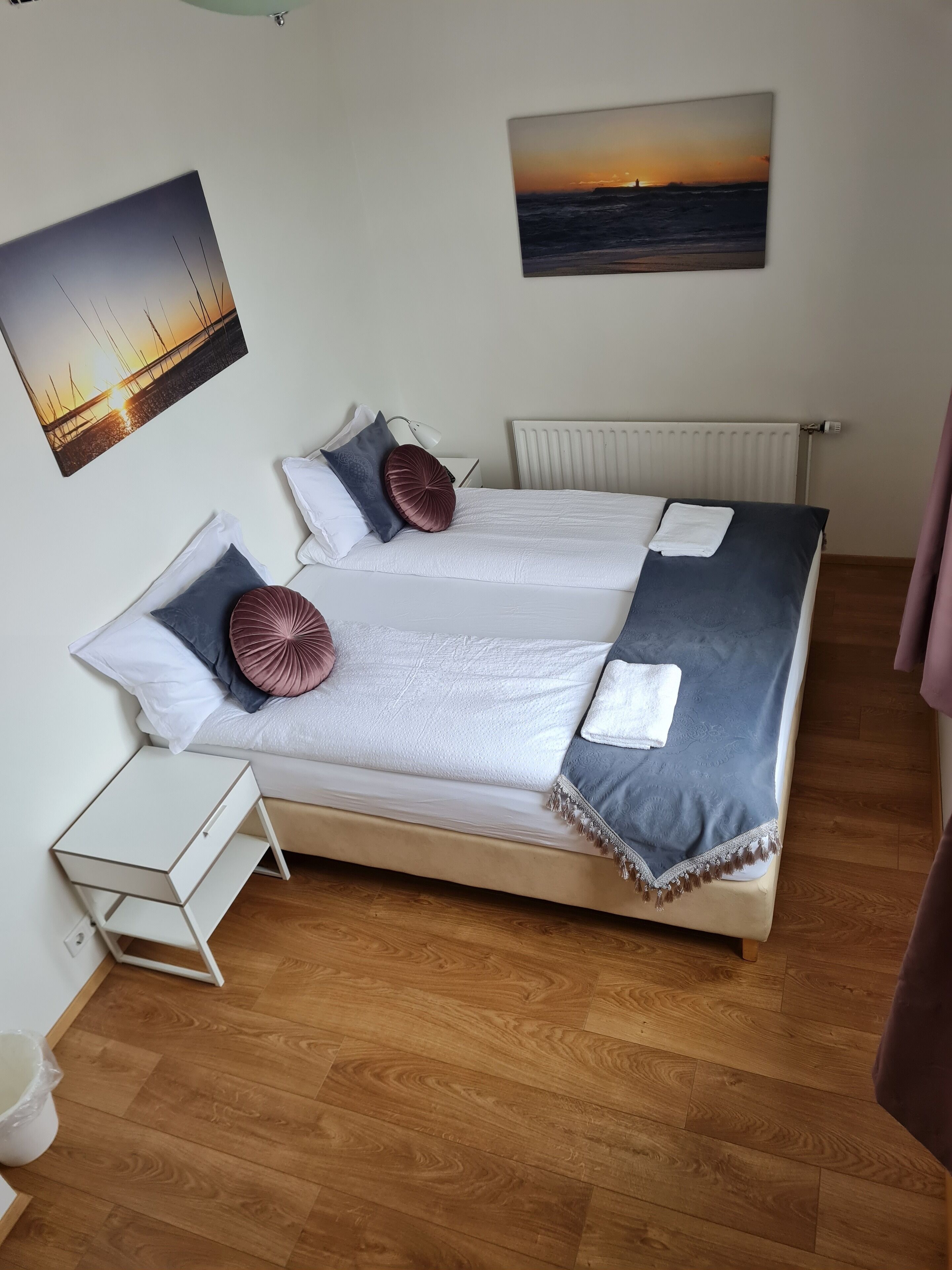 Una cama doble con edredón blanco y gris, dos almohadones decorativos, una mesita de noche blanca y un radiador blanco.