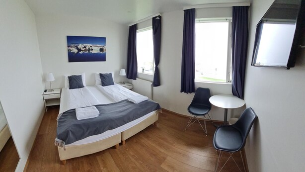 A hotel room with a large bed, two bedside tables, a desk, two chairs, a window with curtains, and a painting on the wall. - Höfn guesthouse (Höfn)