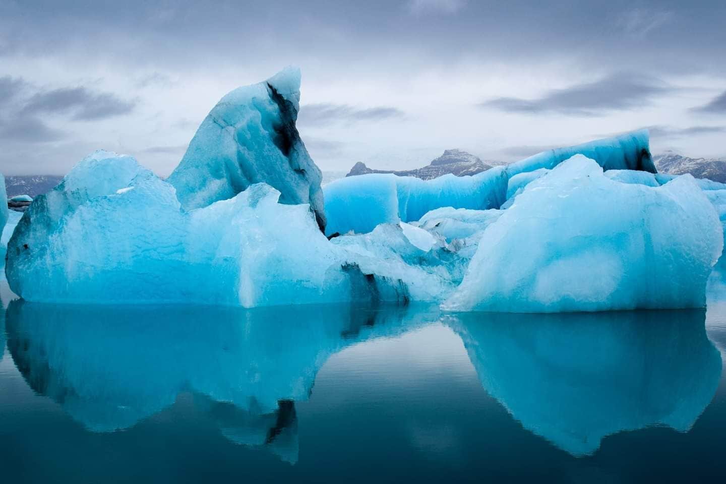 A large iceberg floating in calm, reflective water with a mountainous backdrop.