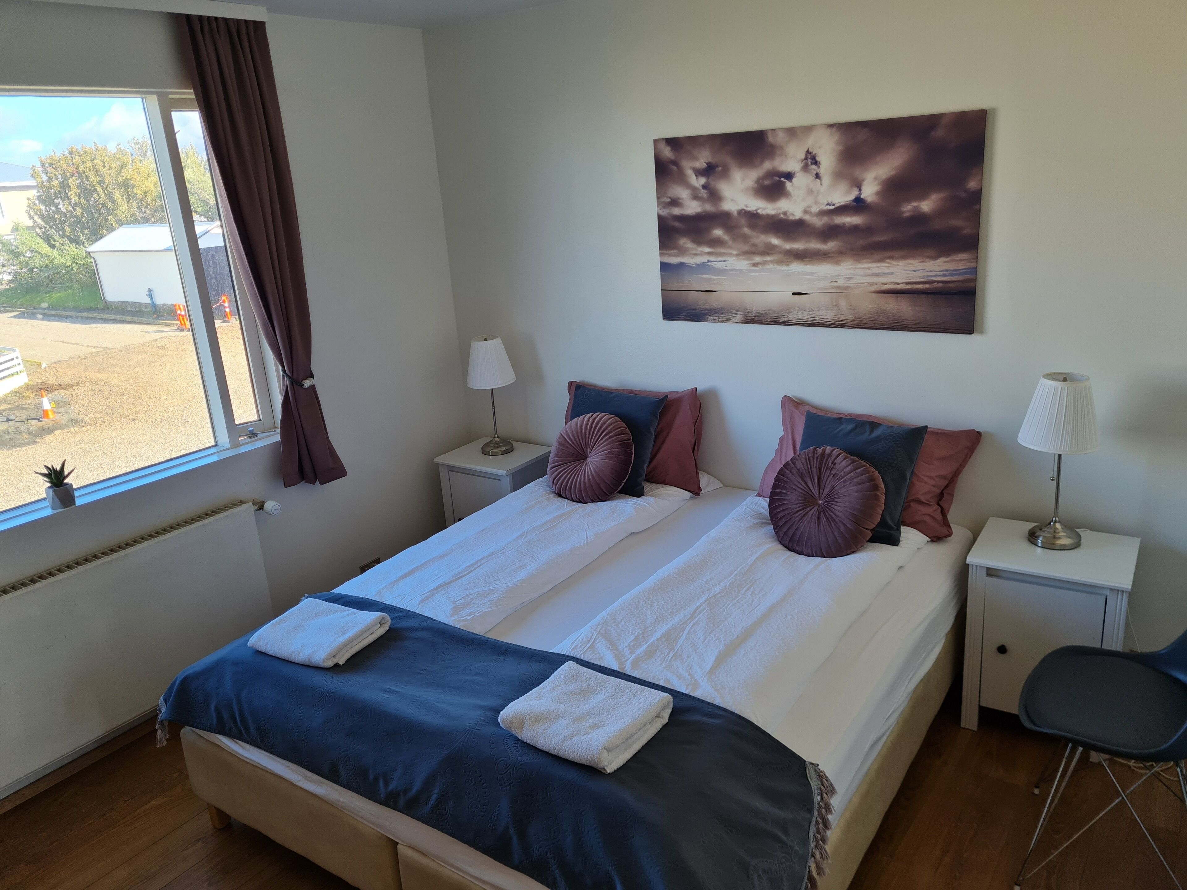 Un lit double avec deux tables de chevet, une fenêtre avec des rideaux, un tableau et une chaise.
