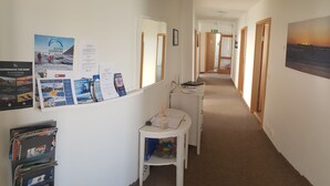 A hotel corridor with a bulletin board displaying travel brochures, a small white table with a lamp, and a bookshelf. - Höfn guesthouse (Höfn)