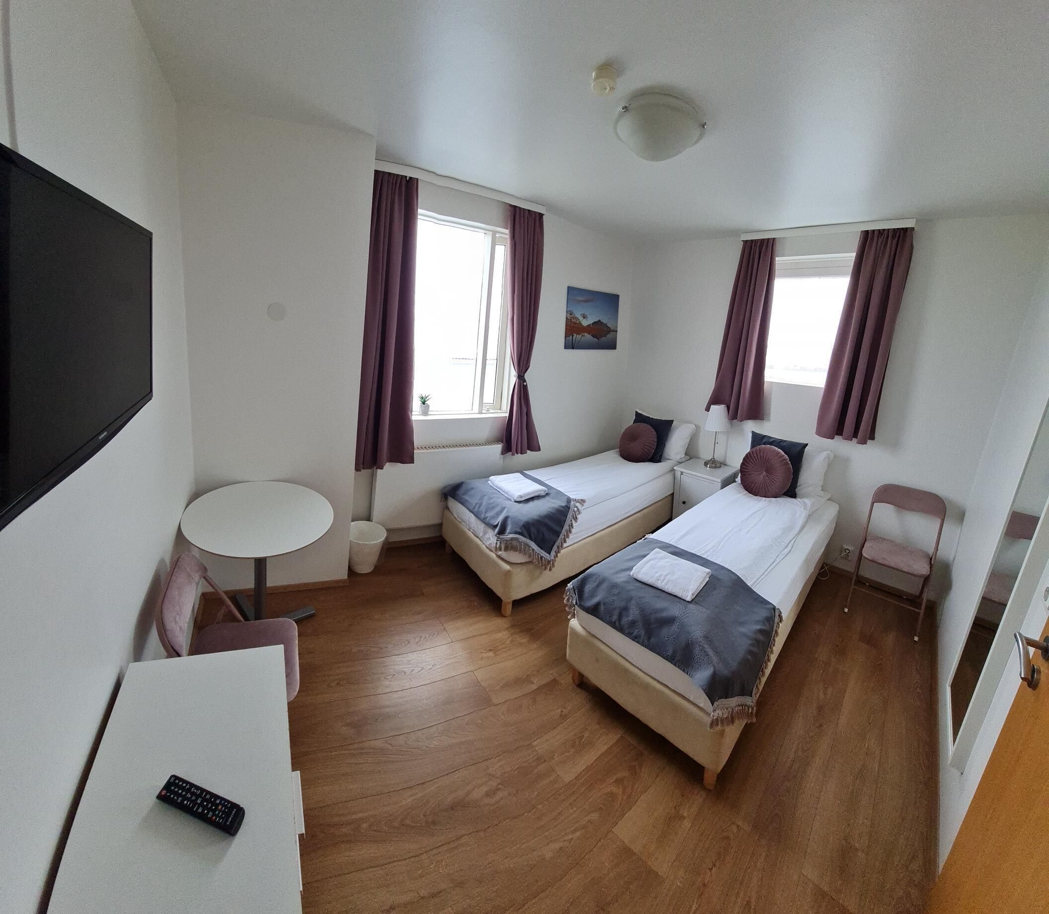 Een kleine hotelkamer met twee eenpersoonsbedden, een tafeltje, een stoel, een televisie en een raam met gordijnen.