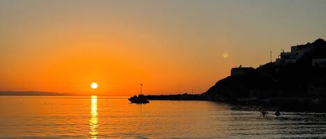 A sunset over a calm sea with a boat and a chair on the shore.