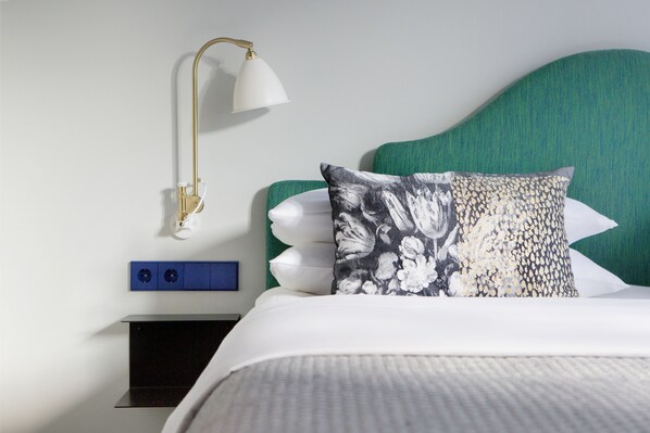 A neatly made bed with a green headboard, a white pillow, and a floral-patterned pillow. There is a white bedside lamp with a gold base and a blue panel below the lamp. - THE FRITZ Düsseldorf (Düsseldorf)