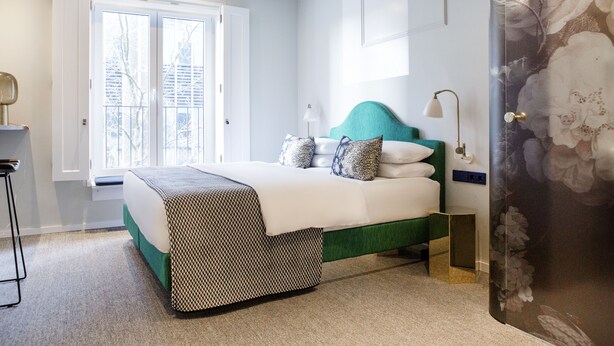 A bedroom with a bed, bedside table, lamp, and a window with a view of trees. - THE FRITZ Düsseldorf (Düsseldorf)
