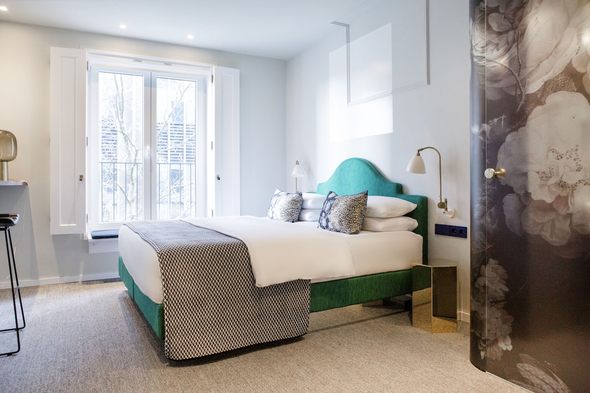 Un dormitorio con cama, mesita de noche, lámpara y ventana con vista a árboles.