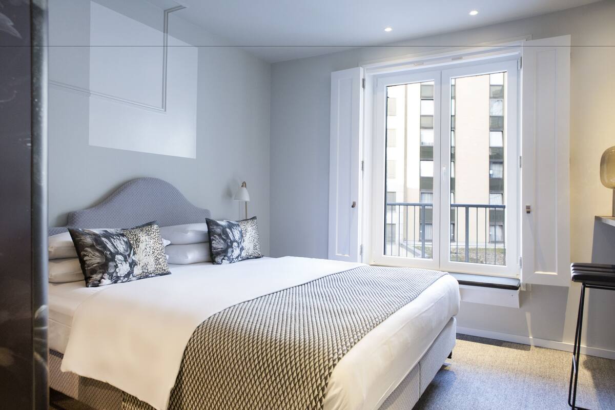 Un dormitorio con cama, mesita de noche, ventana y puerta.